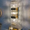 chiếc đèn tường được thiết kế thân đèn làm bằng đồng mạ vàng được cố định trên tường cùng với một hộp đèn có chao đèn là pha lê trong suốt với những đường vân mờ và mềm mại