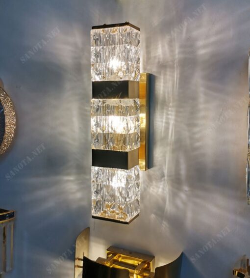 chiếc đèn tường được thiết kế thân đèn làm bằng đồng mạ vàng được cố định trên tường cùng với một hộp đèn có chao đèn là pha lê trong suốt với những đường vân mờ và mềm mại