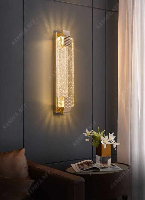 một chiếc đèn gắn tường với thiết kế thân vàng đồng chắc chắn và sang trọng, chao đèn là một tấm pha lê trong suốt có những bong bóng litii tạo hiệu ứng ánh sáng cho chiếc đèn