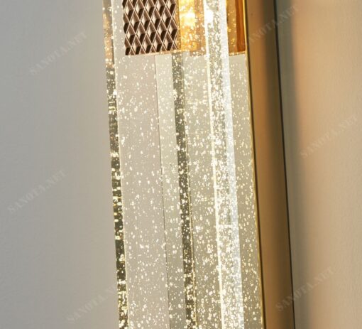 một chiếc đèn gắn tường với thiết kế thân vàng đồng chắc chắn và sang trọng, chao đèn là một tấm pha lê trong suốt có những bong bóng litii tạo hiệu ứng ánh sáng cho chiếc đèn